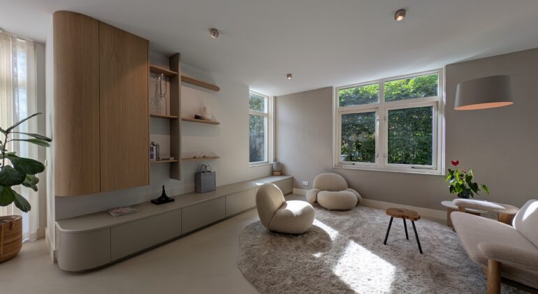 Moderne, minimalistische woonkamer met lichtinval door grote ramen, zachte beige fauteuils, een donzige vloerkleed, een lage wandkast met open planken en een grote kamerplant