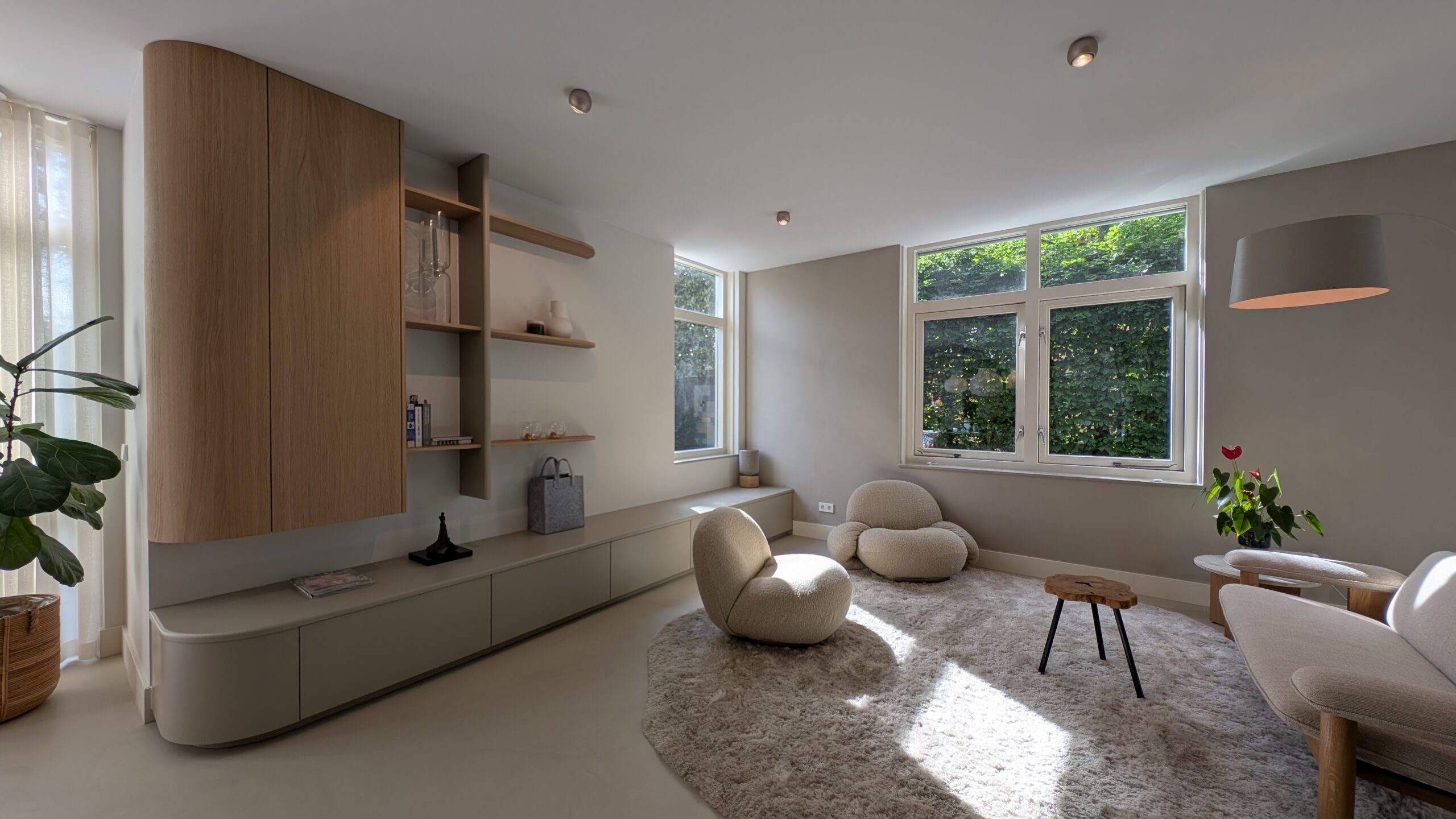 Moderne, minimalistische woonkamer met lichtinval door grote ramen, zachte beige fauteuils, een donzige vloerkleed, een lage wandkast met open planken en een grote kamerplant