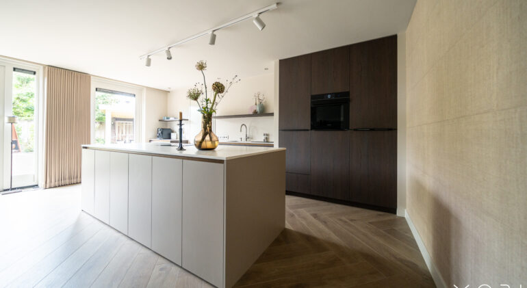 "Een moderne keuken met een groot lichtgrijs keukeneiland en een wand van donkerbruine kasten. Er ligt een houten vloer in visgraatpatroon en er is veel natuurlijk licht dat binnenvalt via de openslaande deuren."