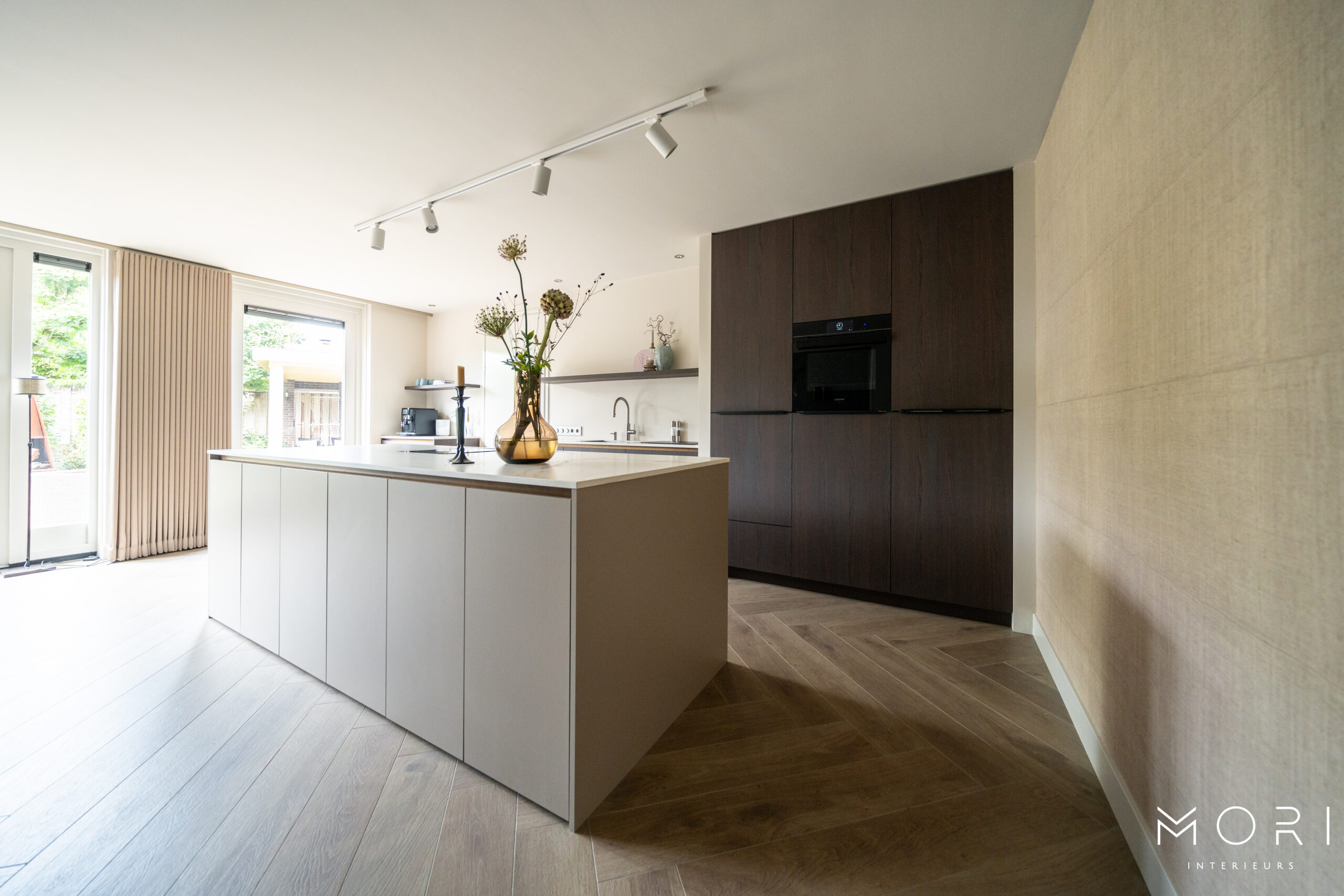 "Een moderne keuken met een groot lichtgrijs keukeneiland en een wand van donkerbruine kasten. Er ligt een houten vloer in visgraatpatroon en er is veel natuurlijk licht dat binnenvalt via de openslaande deuren."